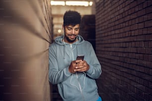 Portrait of happy motivated afro-american young handsome hipster man with earphones standing in the middle of two brick walls while leaning against one and holding mobile.