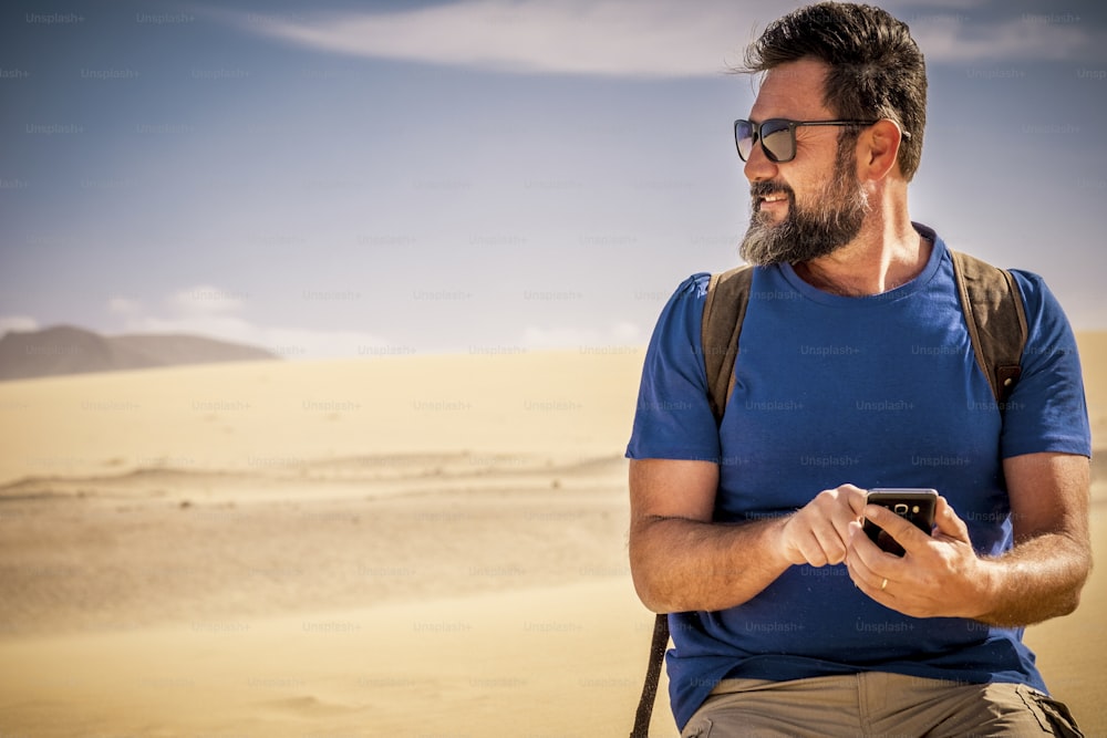 수염 힙스터 스타일을 가진 고위 백인 멋진 남자는 사막 모래 언덕에서 혼자 웃고 야생 야외 장소를 즐기는 인터넷 연결이 가능한 휴대폰을 사용합니다.