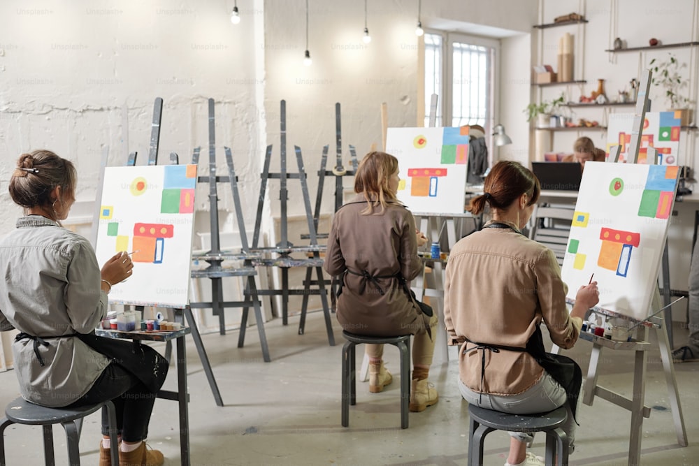 Rear view of several female students of painting class sitting on chairs in front of easels in classroom or studio and working