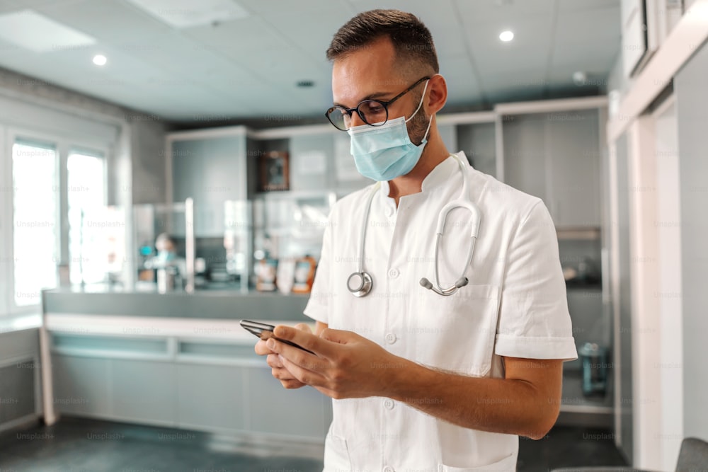 Giovane medico maschio attraente con maschera facciale che utilizza lo smartphone per l'emergenza. Concetto di virus corona.