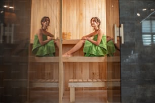 Young and relaxed woman covered with green towel sitting in a small Finnish sauna