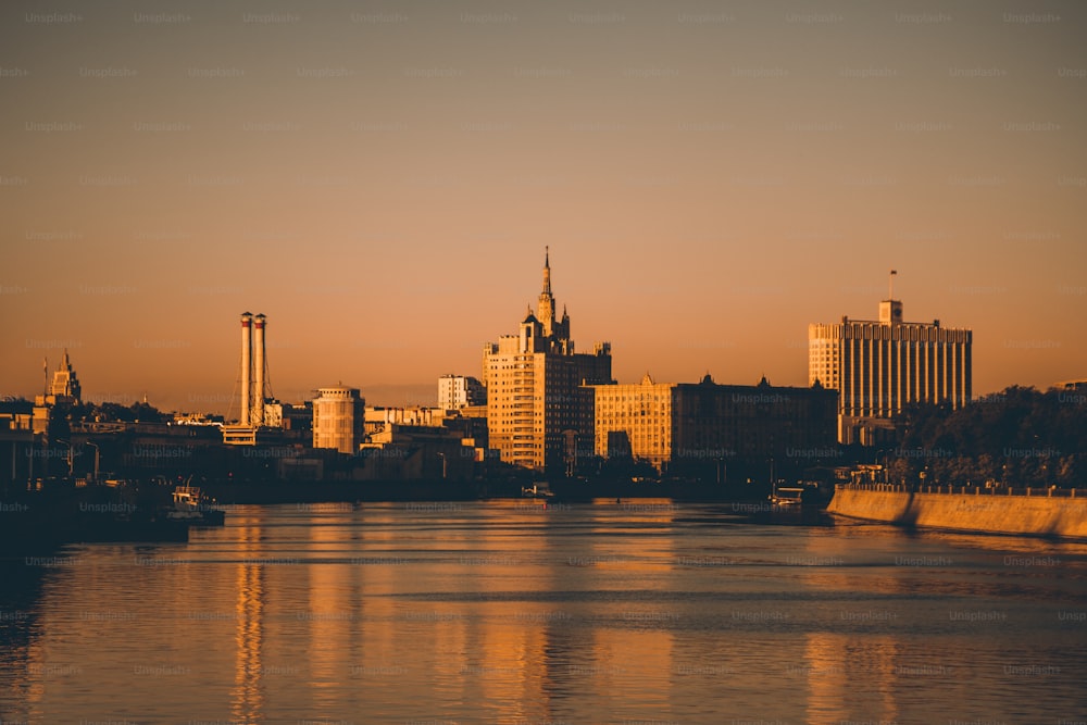 夕暮れ時のロシア・モスクワの街並み:前景にモスクワ川、ホワイトハウスのシルエット、尖塔のある高層ビル、2本の煙突、オレンジ色の夕日に照らされた他の建物