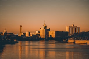 Cityscape of Moscow, Russia during the sunset: Moscow river in the foreground, silhouettes of the White House, the high-rise with the spire, two chimneys and other buildings lit by orange evening sun