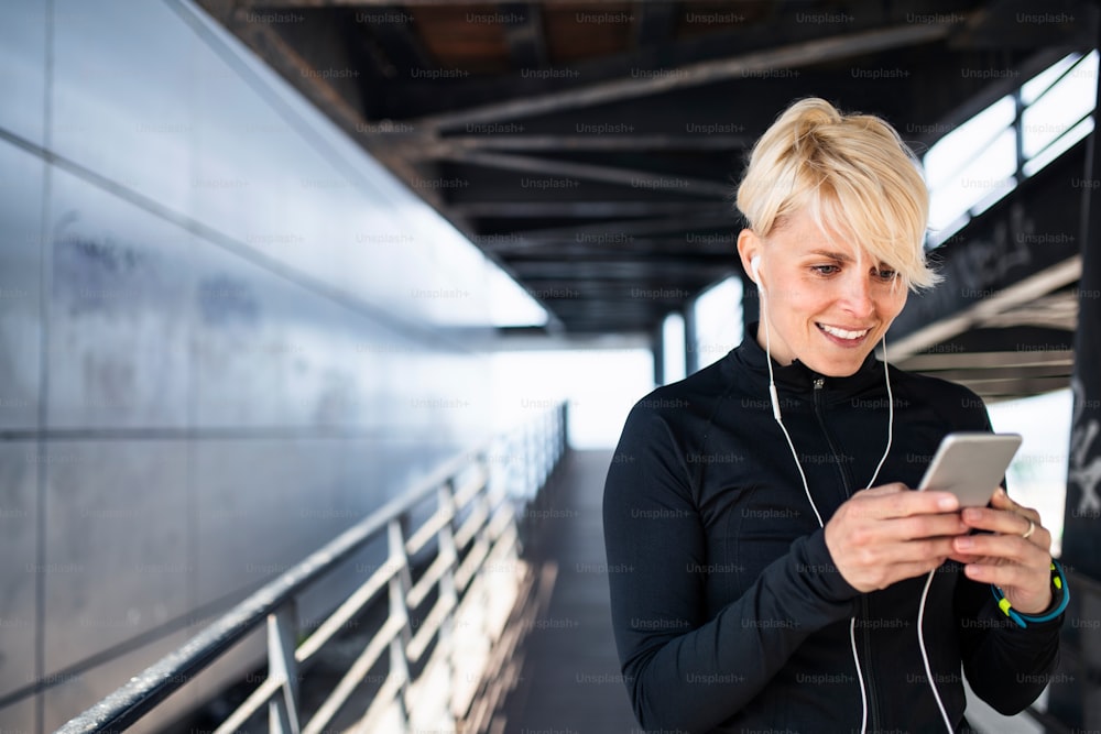 A young sportswoman with earphones and smartphone doing exercise outdoors in city, resting.