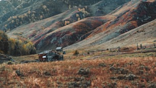Incrível cenário montanhoso com uma cabana de madeira abandonada e o prado em primeiro plano e cume de colinas e com pastagens e barracas de edifícios ao fundo; início do outono nas montanhas de Altai