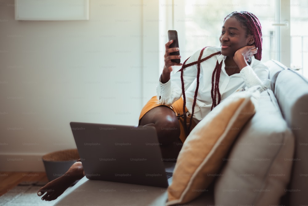 An elegant young cheerful black woman with long braids is using her smartphone to reply a message while working at home: sitting barefooted on the sofa with cushions and her laptop in front of her