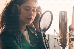 Portrait of beautiful curly woman in leather jacket and hat recording vocals in music studio on professional sound equipment, wearing headphones