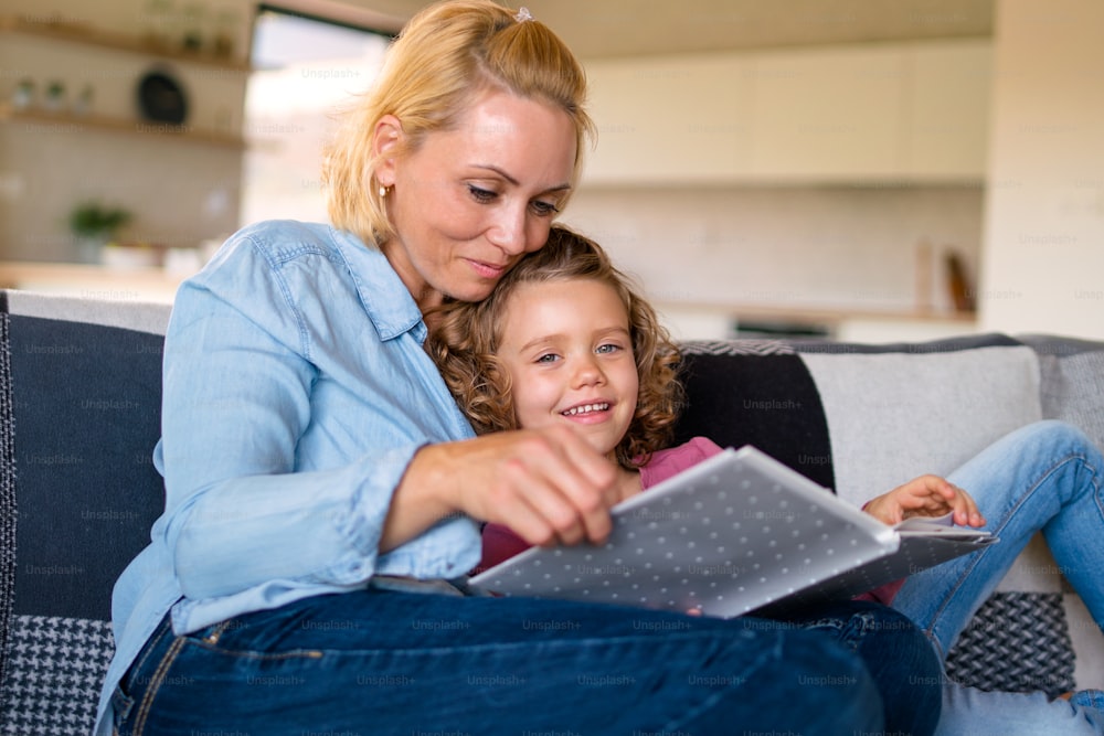 Une jolie petite fille avec sa mère assise sur un canapé à l’intérieur à la maison, lisant un livre d’histoires.