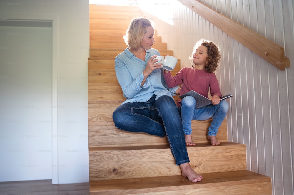 Una vista frontal de una linda niña pequeña con su madre en el interior de la casa, sentada en la escalera.