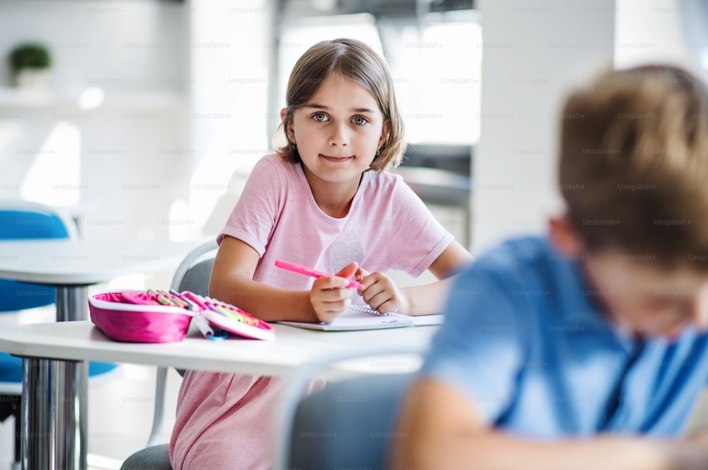Ein Porträt eines kleinen, glücklichen Schulmädchens, das am Schreibtisch im Klassenzimmer sitzt und in die Kamera schaut.