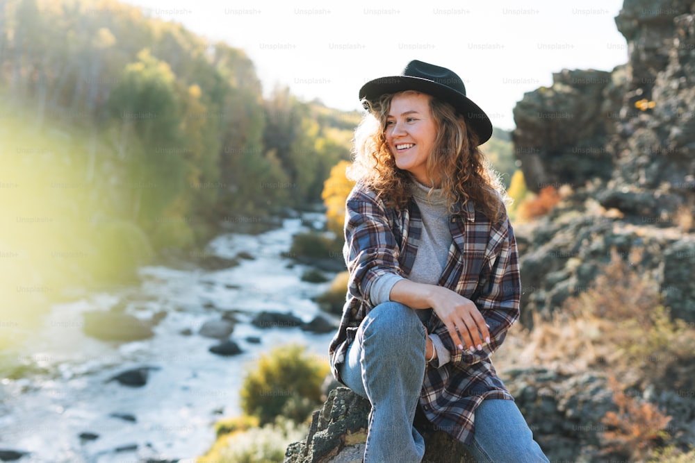 Young beautiful woman with curly hair in felt hat and plaid shirt in jeans looks at view of mountains and river, hiking on autumn nature, sustainable environment