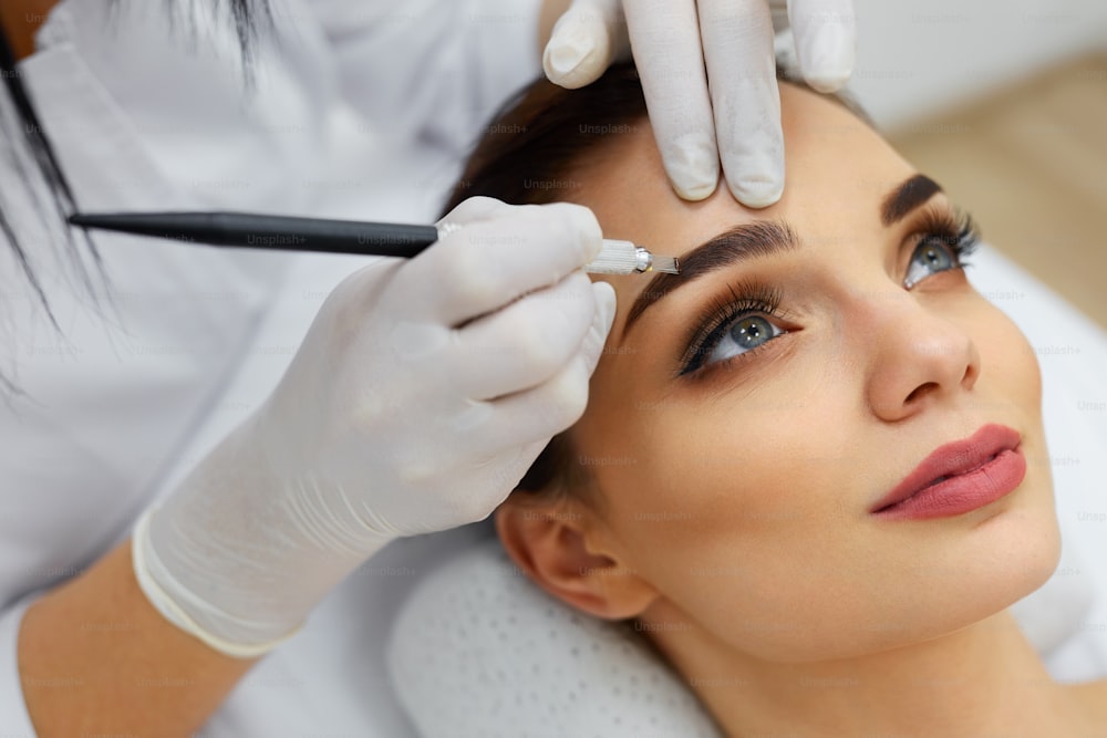 Maquillaje. Manos De Esteticista Haciendo Tatuaje De Cejas En La Cara De La Mujer.Maquillaje Permanente De Cejas En El Salón De Belleza. Primer Plano De Especialista Haciendo Tatuación De Cejas Para Mujer. Tratamiento de Cosmetología. Alta resolución