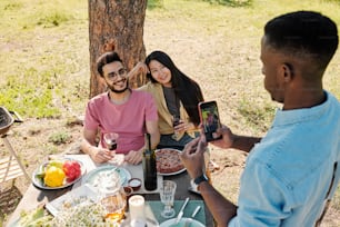 Giovane uomo africano con smartphone che fotografa la coppia interculturale che si siede sotto il pino davanti al tavolo servito per la cena
