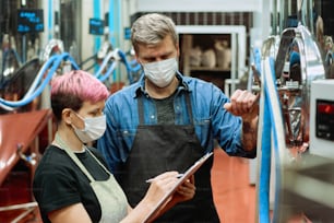 Two young brewers in workwear and protective masks discussing notes in clipboard while blond female pointing at document during work