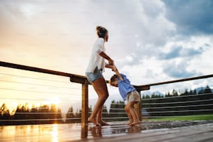 Happy mother with small daughter playing in rain on patio of wooden cabin, holiday in nature concept.