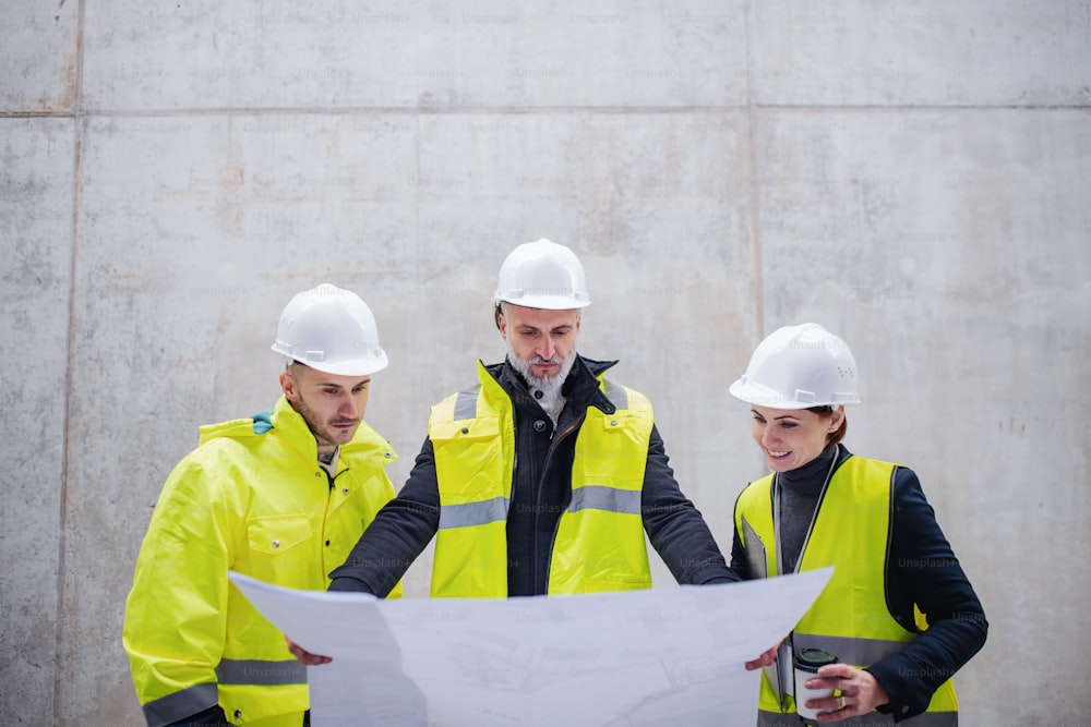 Un grupo de ingenieros de pie contra un muro de hormigón en una obra de construcción, sosteniendo planos.