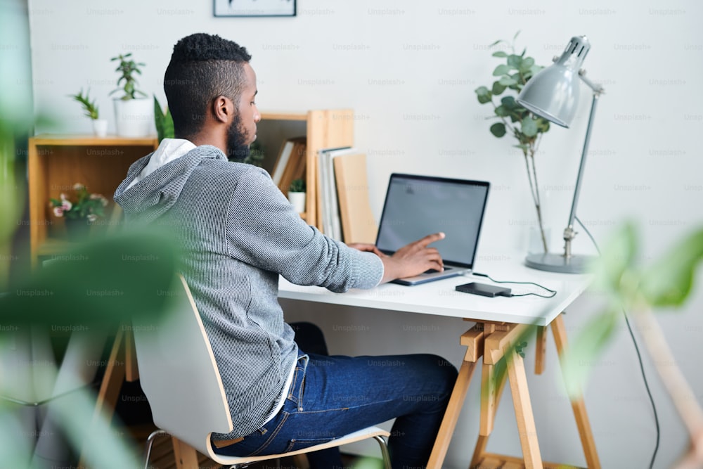 Junger interkultureller Mann in Casualwear bereitet ein neues Geschäftsprojekt vor und nimmt Ideen aus dem Netz, während er vor dem Laptop sitzt