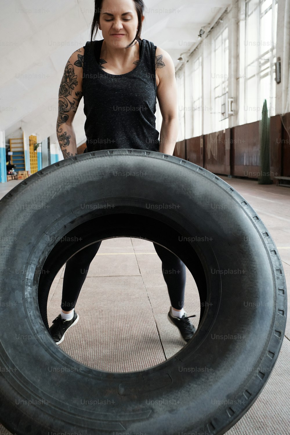 Femme athlétique déterminée avec des bras tatoués retournant un gros pneu lourd tout en s’entraînant dans un gymnase