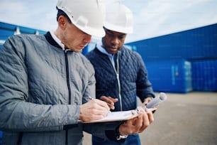 Two engineers standing by shipping containers on a large commercial freight dock discussing an inventory list on a clipboard