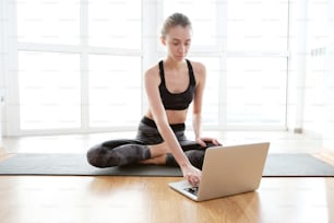 Full body shot of young skinny woman using laptop while sitting on mat in front of big bright window. She is wearing tank and dark pants