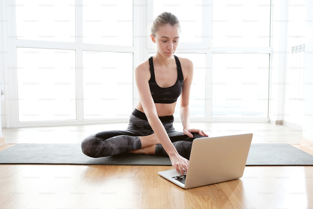 Full body shot of young skinny woman using laptop while sitting on mat in front of big bright window. She is wearing tank and dark pants