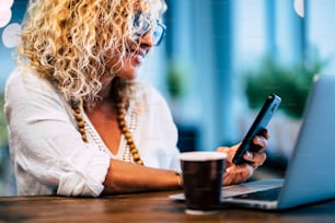 close up of woman smiling and looking at her phone with her laptop and coffee - beautiful and worker adult lady using and working with modern devices -   office workstation technology devices