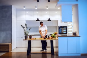Toute la longueur d’un chef caucasien souriant en uniforme blanc hachant l’oignon et préparant le repas. Sur le comptoir de la cuisine, il y a des légumes et des épices.