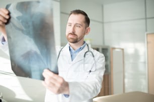 Young radiologist in uniform looking at x-ray image before making medical diagnosis