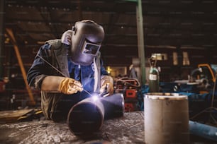 Welder in protective uniform and mask welding metal pipe on the industrial table while sparks flying.