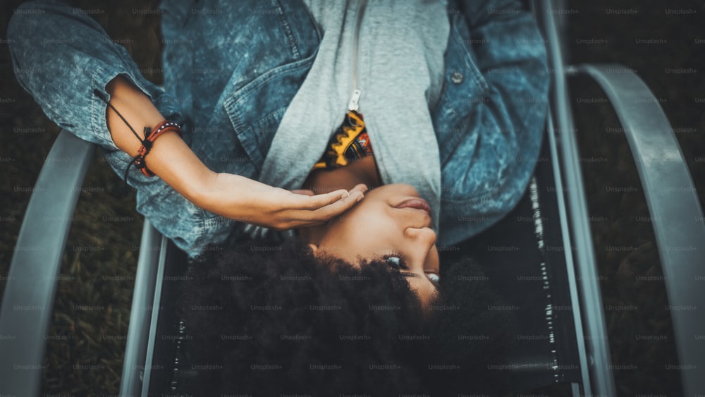Encantadora menina afro-americana está deitada ao ar livre na poltrona reclinável e olhando para o lado; menina negra com cabelos afro encaracolados na espreguiçadeira do lado de fora; fêmea brasileira adulta pensativa em uma chaise-longue de rua