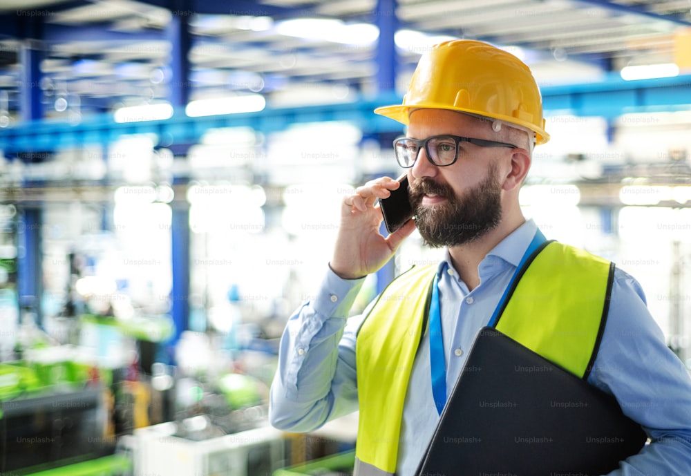 Vue latérale d’un technicien ou d’un ingénieur portant un casque de sécurité debout dans une usine industrielle, à l’aide d’un téléphone.