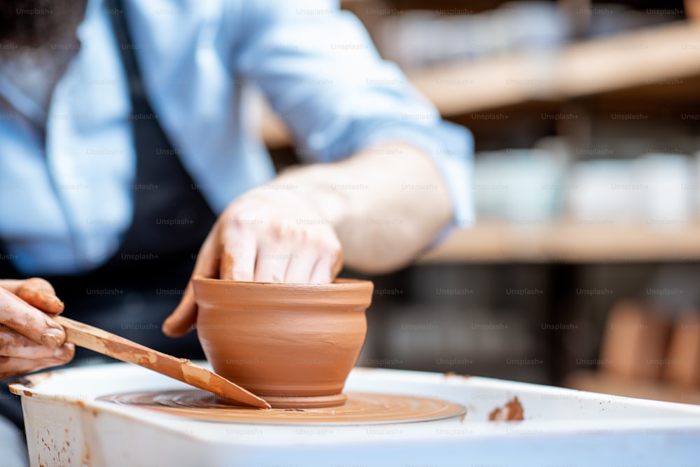 Man making clay jug forming shape by hands on the pottery wheel indoors, close-up view