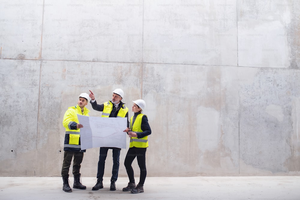 Un gruppo di ingegneri in piedi contro un muro di cemento in cantiere, con in mano dei progetti. Copia spazio.