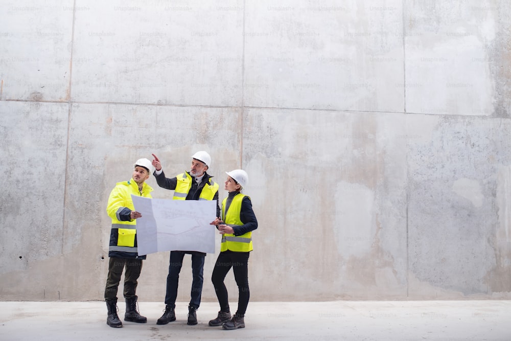 Un grupo de ingenieros de pie contra un muro de hormigón en una obra de construcción, sosteniendo planos. Espacio de copia.