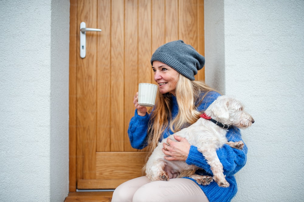Mature woman relaxing outdoors by front door at home with coffee and pet dog. Copy space.