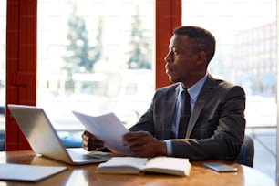 Serious African businessman in formalwear working with papers while sitting by table in office