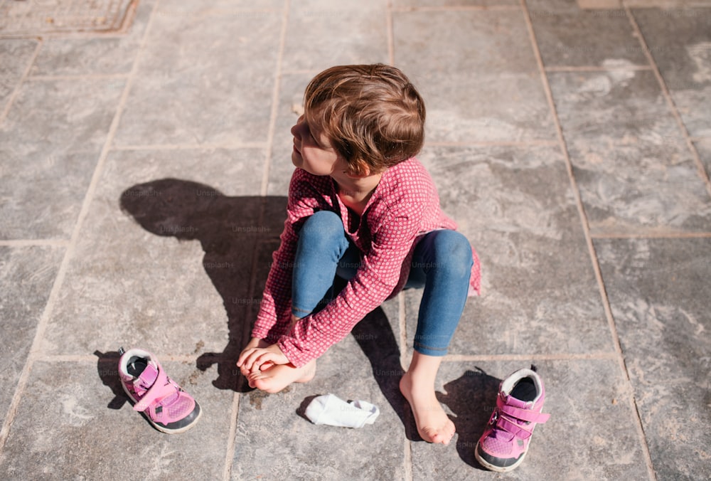 A top view of small girl sitting outdoors in town on pavement, taking off shoes.