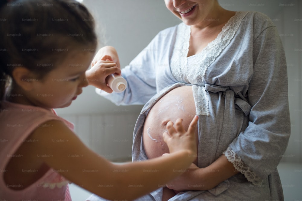 Small girl creaming belly of unrecognizable pregnant mother indoors in bathroom at home.