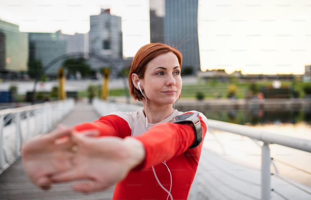 Vista frontal de una joven corredora con auriculares en la ciudad, estirándose en el puente.
