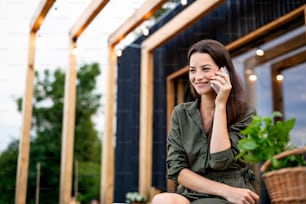 Happy young woman with smartphone outdoors, weekend away in container house in countryside.