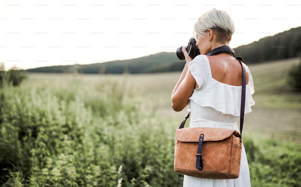 A rear view of woman photographer with a leather bag and camera in nature, taking photographs. Copy space.
