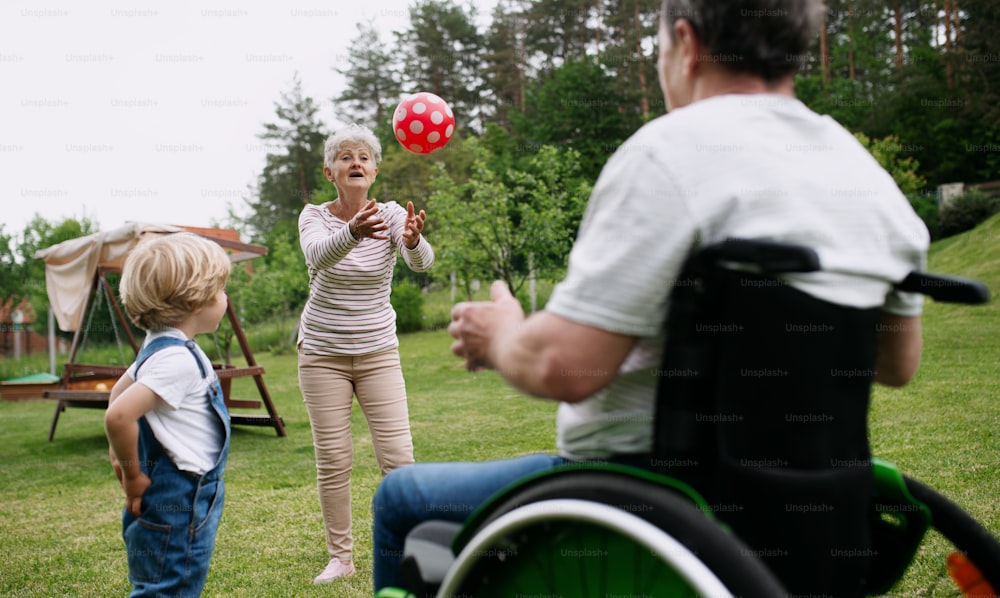 Happy small boy with senior grandparents in wheelchair playing with a ball in garden.