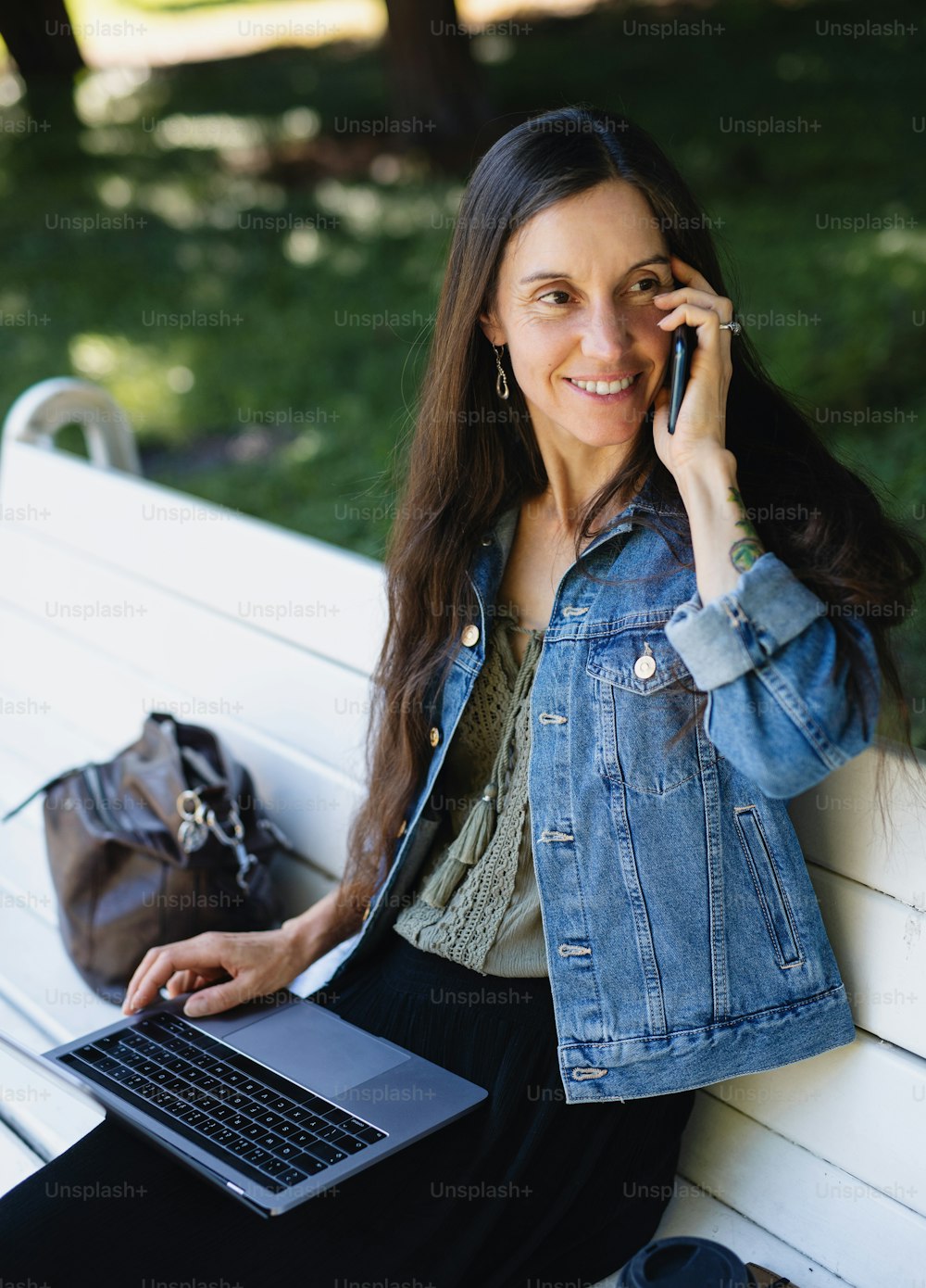 Mature woman with laptop and smartphone sitting on bench outdoors in city or town park, working.
