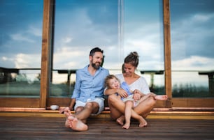 Happy family with small daughter sitting on patio of wooden cabin, holiday in nature concept.