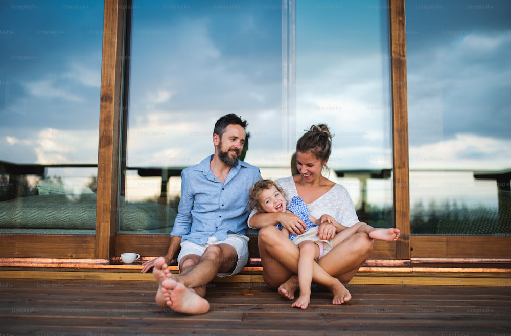Happy family with small daughter sitting on patio of wooden cabin, holiday in nature concept.