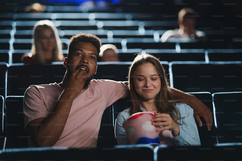 A front view of cheerful young couple with popcorn in the cinema, watching film.
