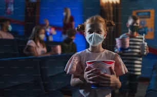 A happy small child with popcorn standing and looking at camera in the cinema, coronavirus concept.