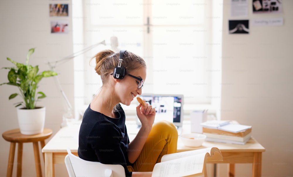 Una giovane studentessa universitaria felice seduta al tavolo di casa, che usa cuffie e laptop durante lo studio.
