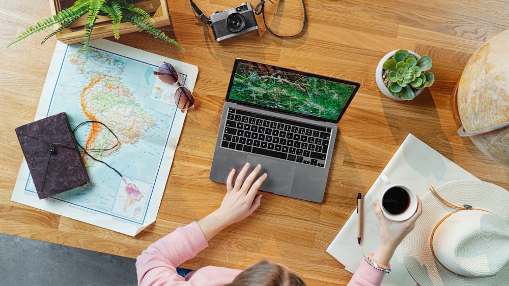 Vista dall'alto di giovane donna irriconoscibile con laptop e mappe che pianificano le vacanze di viaggio, concetto di viaggio desktop.
