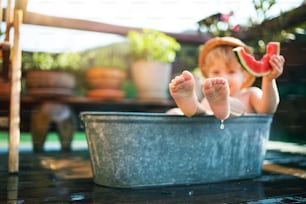 Happy small boy with a hat in bath tub outdoors in garden in summer, eating watermelon.